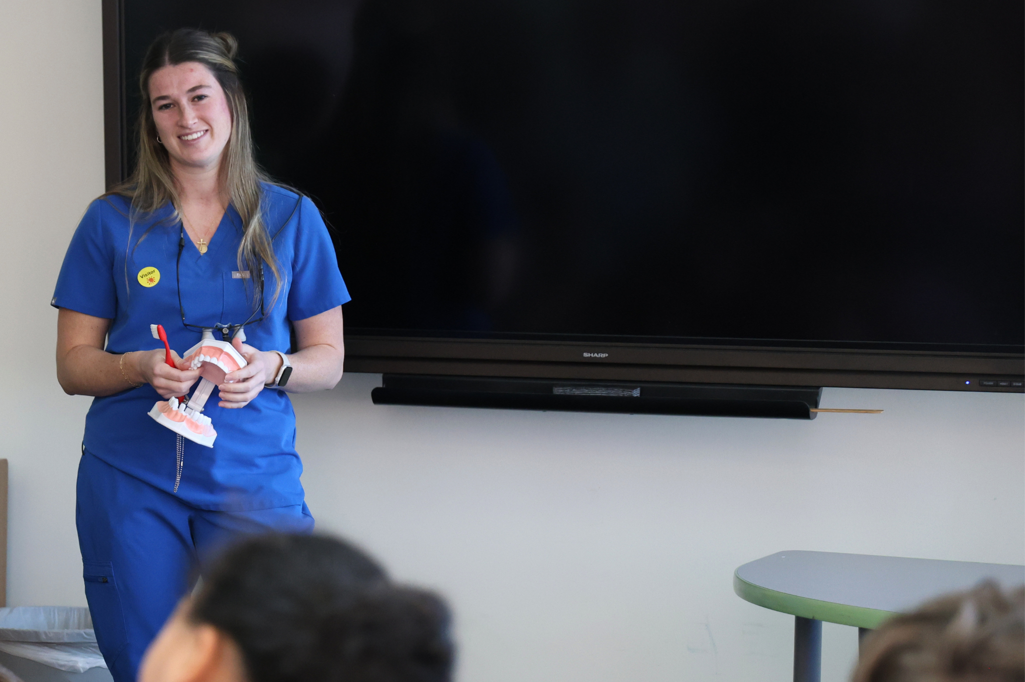 A woman in scrubs stands in front of a group of students holding a model of teeth and a toothbrush